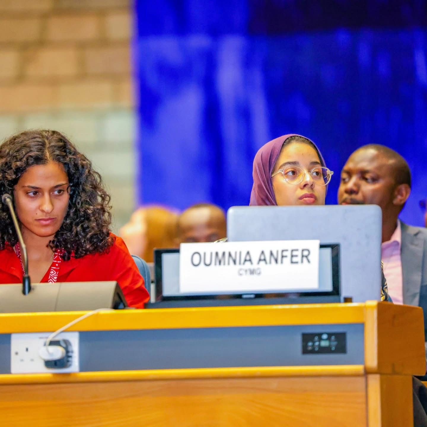 Oumnia wearing a mauve hijab at a conference desk with her CYMG nameplate